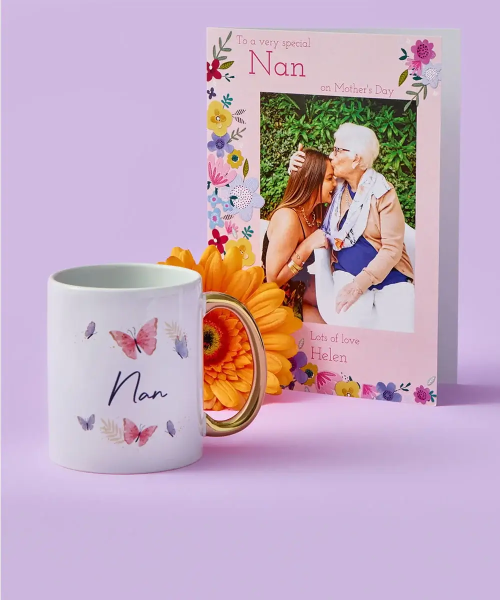 A floral Nan Mother’s Day card featuring a warm moment between two people is displayed behind a white mug decorated with pastel butterflies and the word Nan, with an orange flower placed in front on a pastel purple background.